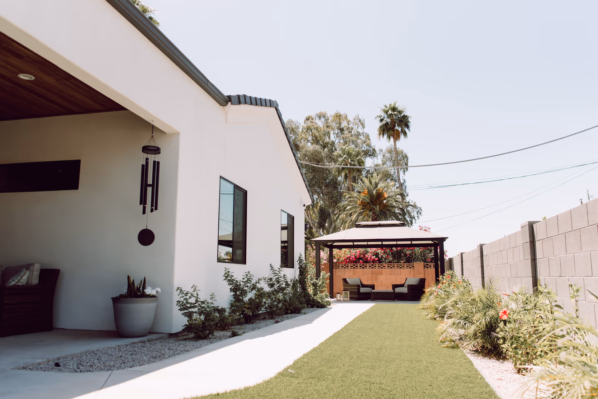 Outdoor view of a garden area beside a white building with a covered patio on the left and a gazebo with seating at the end of a green lawn. There are plants and flowers along the pathway and a tall palm tree in the background under a clear sky.