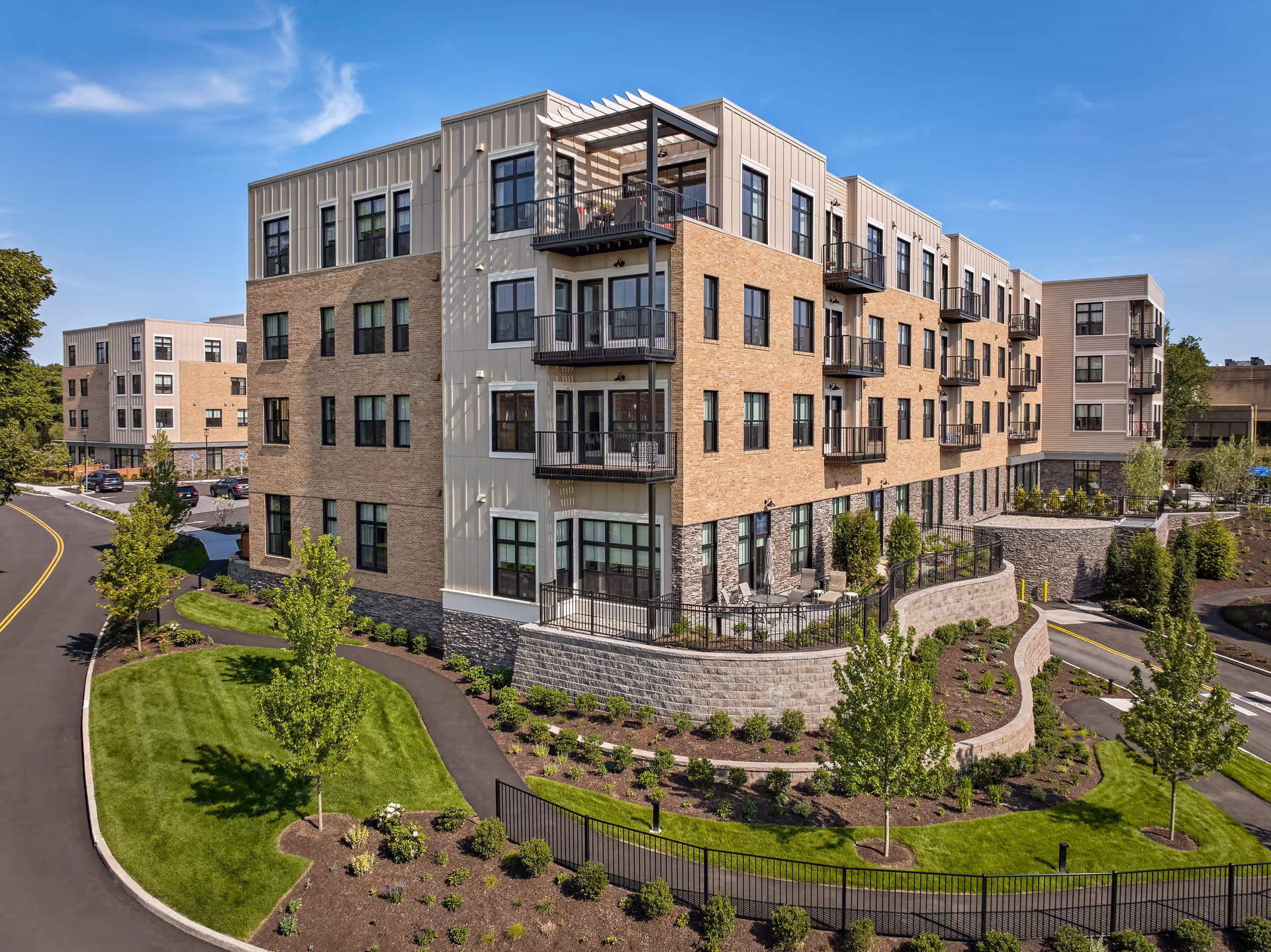 Exterior view of a modern multi-story senior living facility with balconies, landscaped grounds, trees, and a paved road under a clear blue sky.
