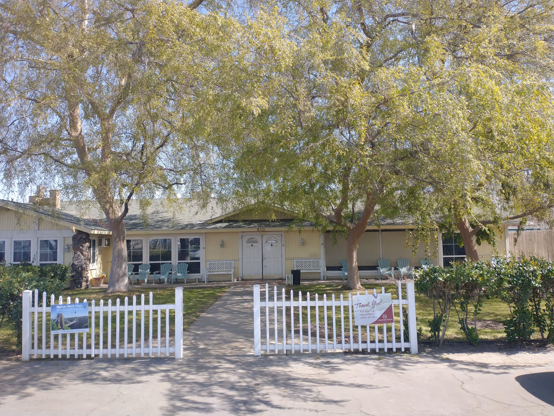 Front view of a single-story assisted living building with a white picket fence, large trees, and chairs on the porch.