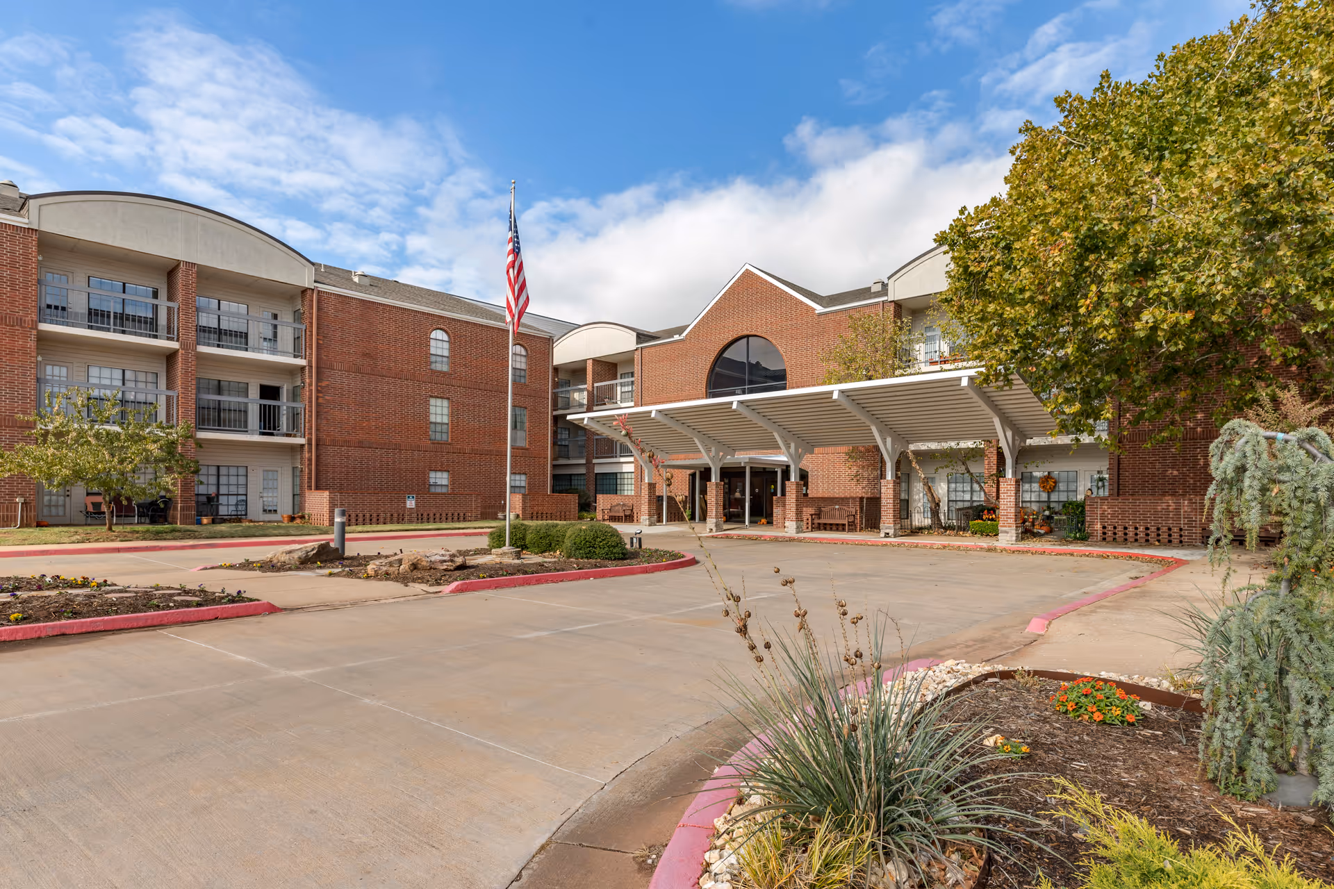 Brick senior living facility exterior with a covered entrance and flag, parking lot, and landscaping.
