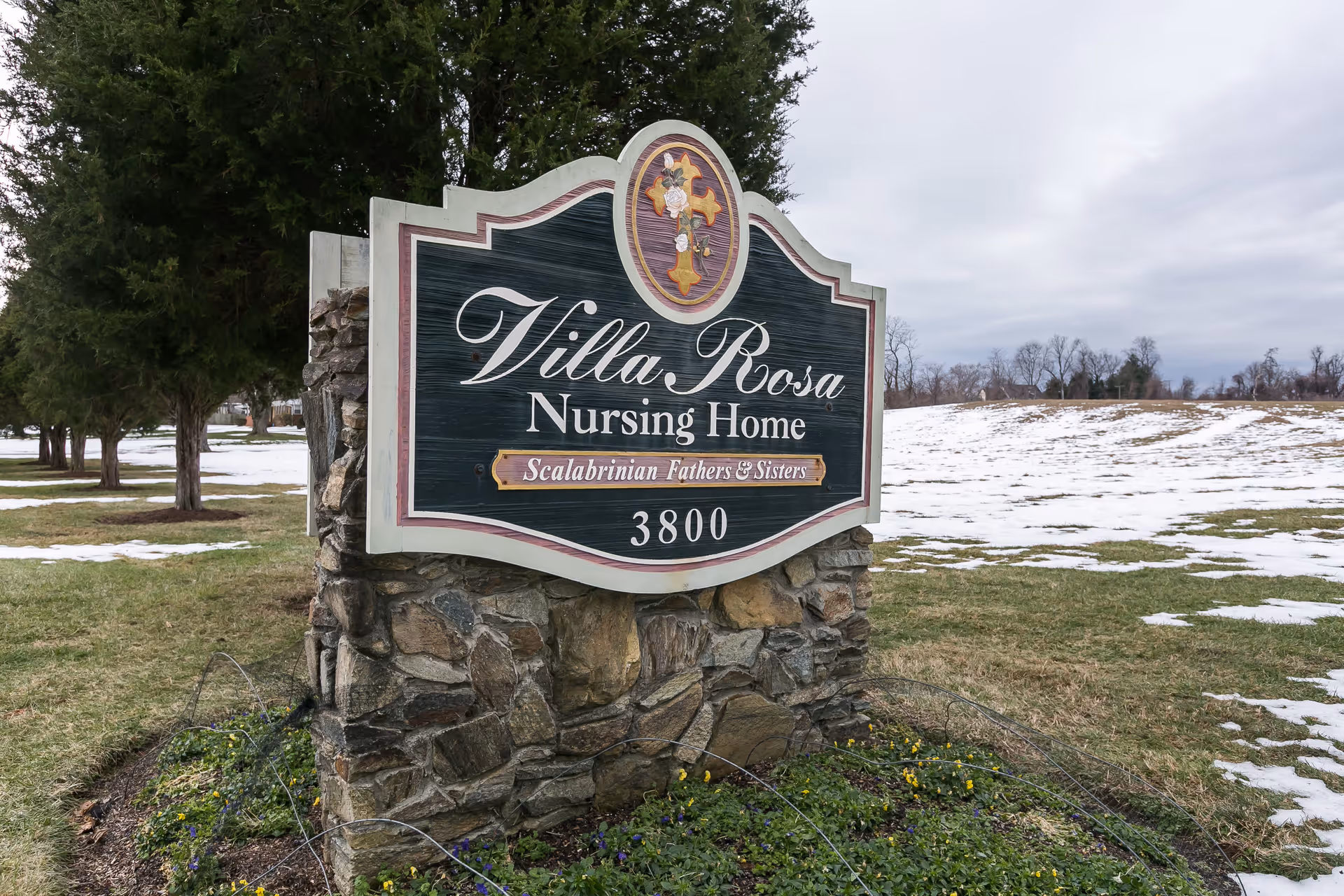 A stone and wood sign for Villa Rosa Nursing Home with the text 'Scalabrinian Fathers & Sisters' and the number 3800, set in a grassy area with patches of snow and trees in the background under a cloudy sky.