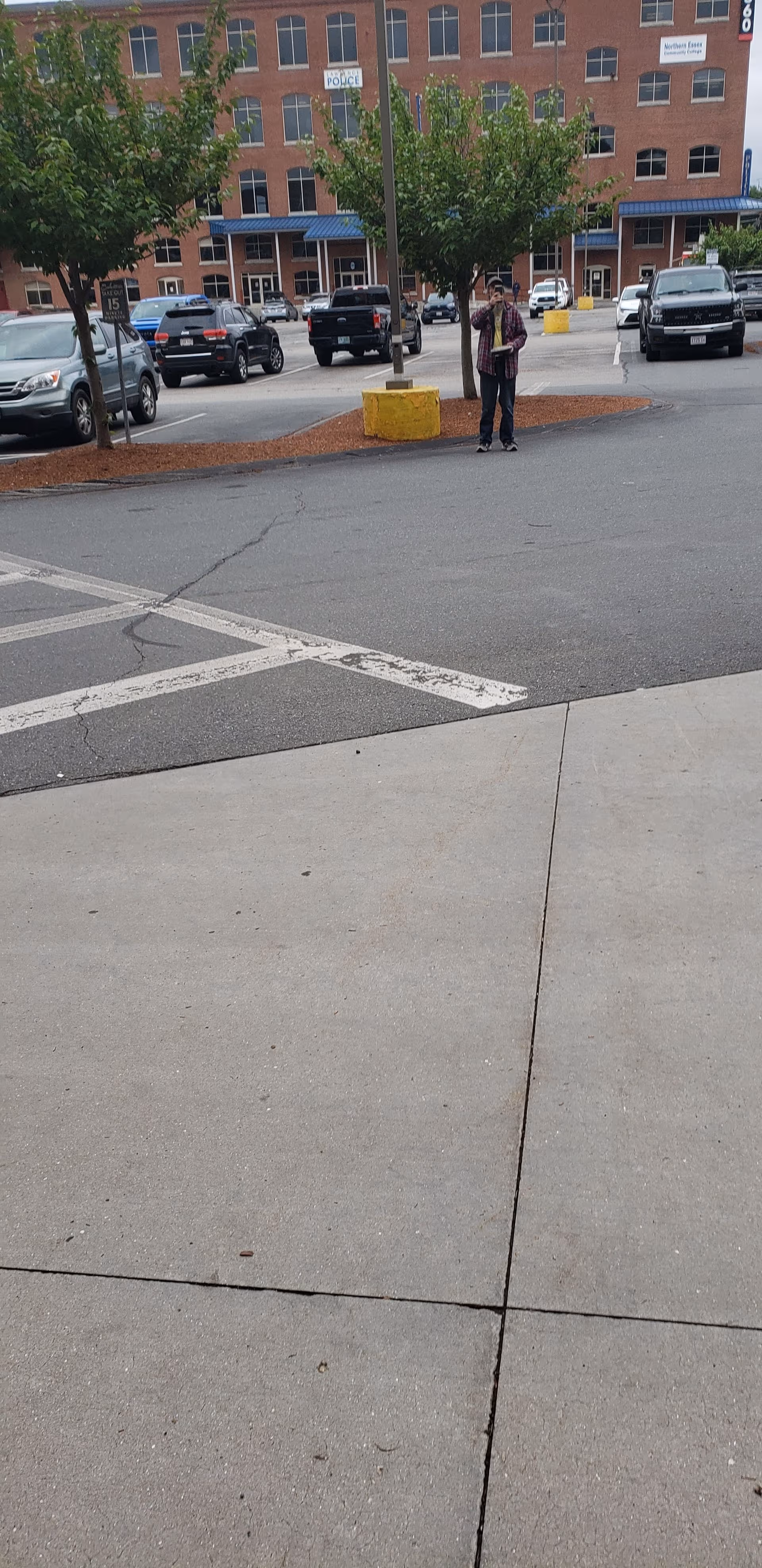 A person stands near a yellow barrier in a parking lot in front of brick building.