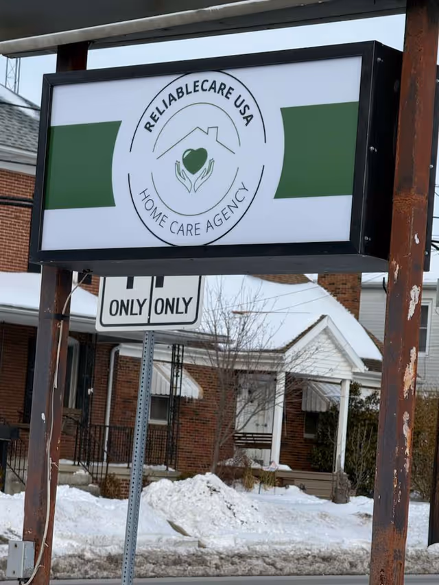 Outdoor sign for Reliablecare USA Home Care Agency with a brick building in a snowy scene