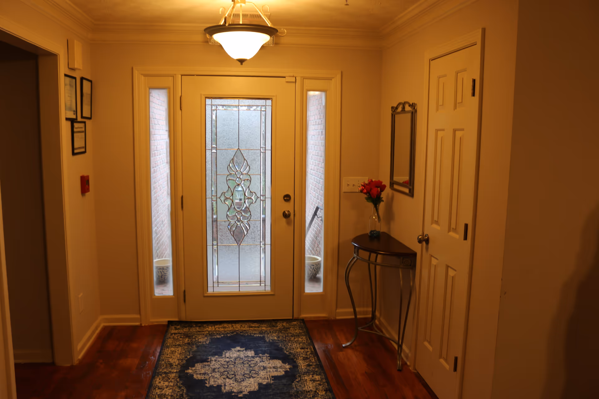 Entryway of a facility with a decorative glass front door flanked by two narrow windows. There is a small table with a vase of red flowers and a mirror above it on the right side. The floor has a blue patterned rug, and the walls are painted beige with framed certificates on the left wall.