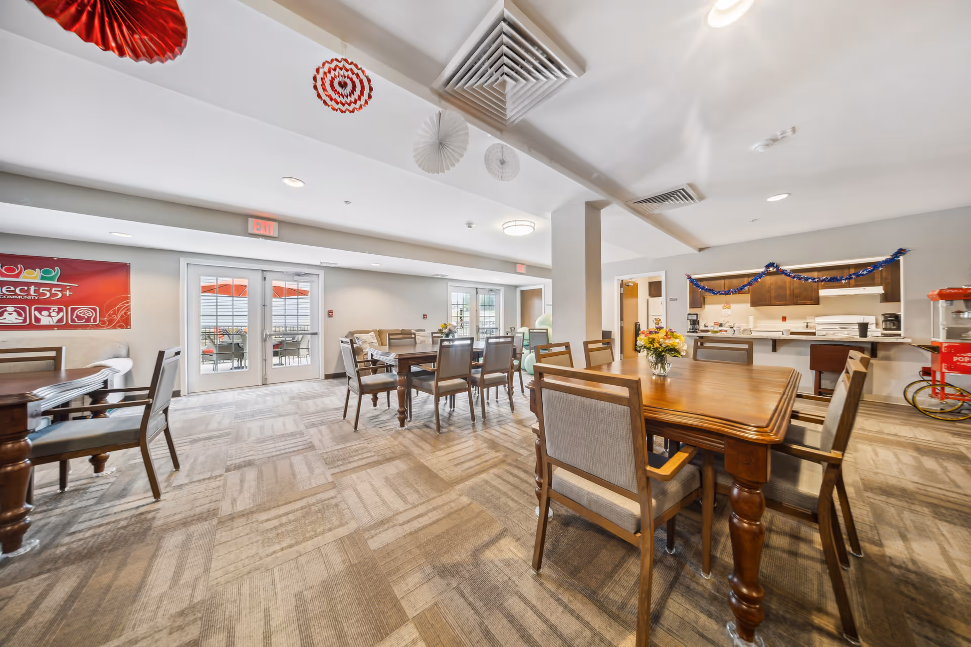 Interior view of a bright and spacious common dining area in a retirement community. The room features several wooden tables with cushioned chairs arranged around them. There are decorations hanging from the ceiling and a popcorn machine in the corner. Large glass doors lead to an outdoor patio area with red umbrellas. The kitchen area is visible in the background with cabinets and appliances.