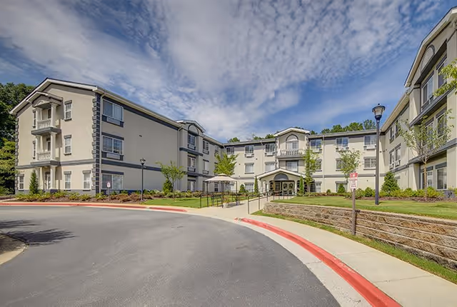 Exterior view of a three-story senior living facility building with beige walls and dark trim, surrounded by landscaped greenery and a curved driveway with red curbs under a partly cloudy sky.