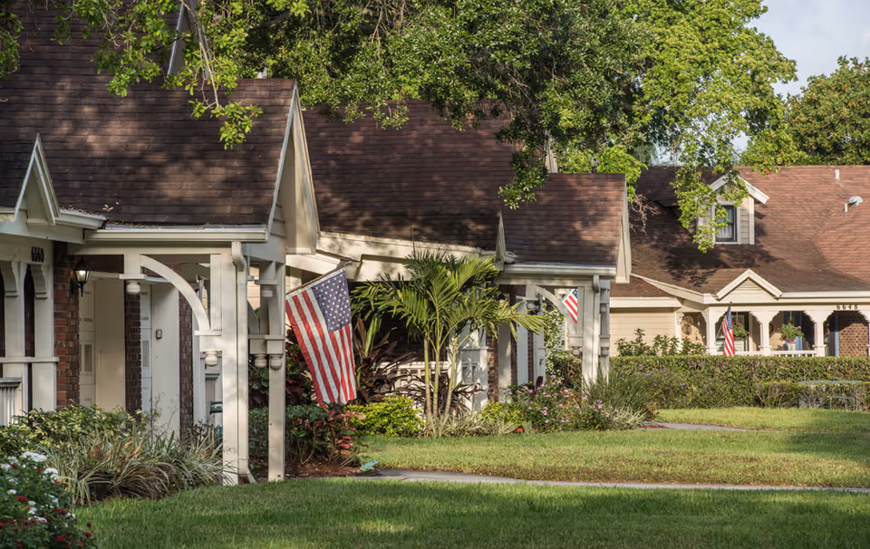 Row of single-story cottage-style residences with porches, American flags, and landscaped lawns under large trees.