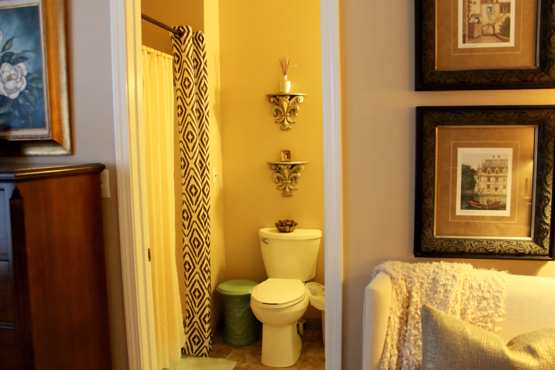 View of a small bathroom with a white toilet, a green trash bin, and a shower with a patterned curtain. The bathroom is adjacent to a living area with a white couch draped with a textured blanket and two framed pictures on the wall.