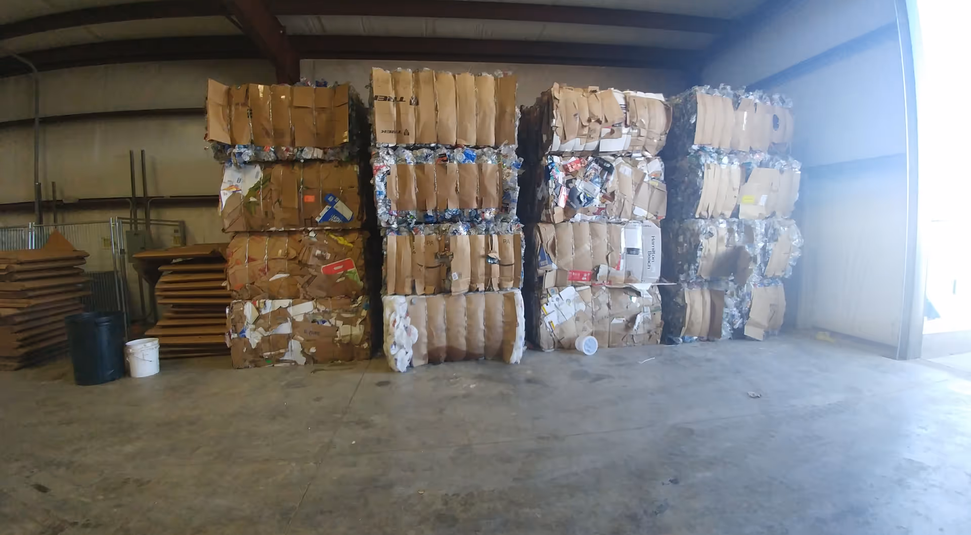 Inside a warehouse, tall stacks of cardboard recycling bales fill the space.