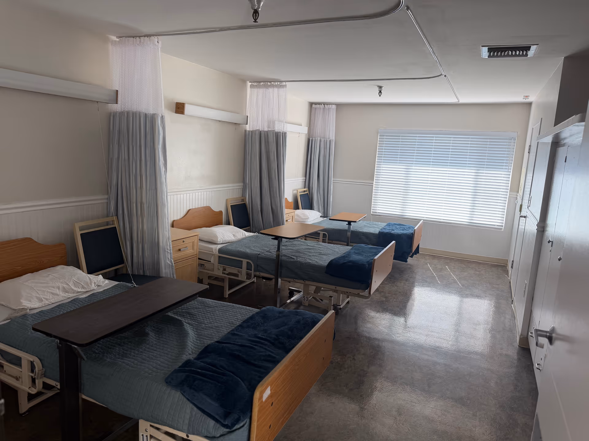 A ward-style hospital room with multiple beds, curtains, and a large window.