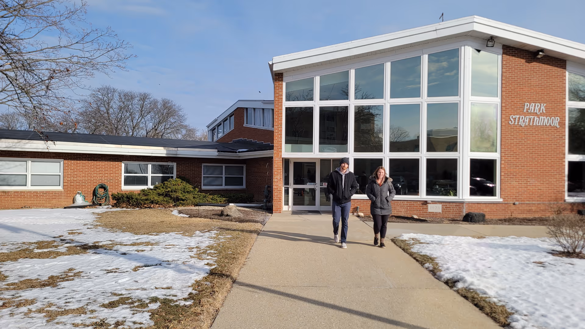 Two people walk toward a brick senior living facility entrance with large windows.