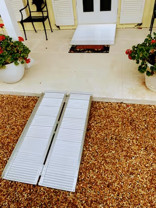 A portable metal wheelchair ramp leads to a front door with potted plants nearby.