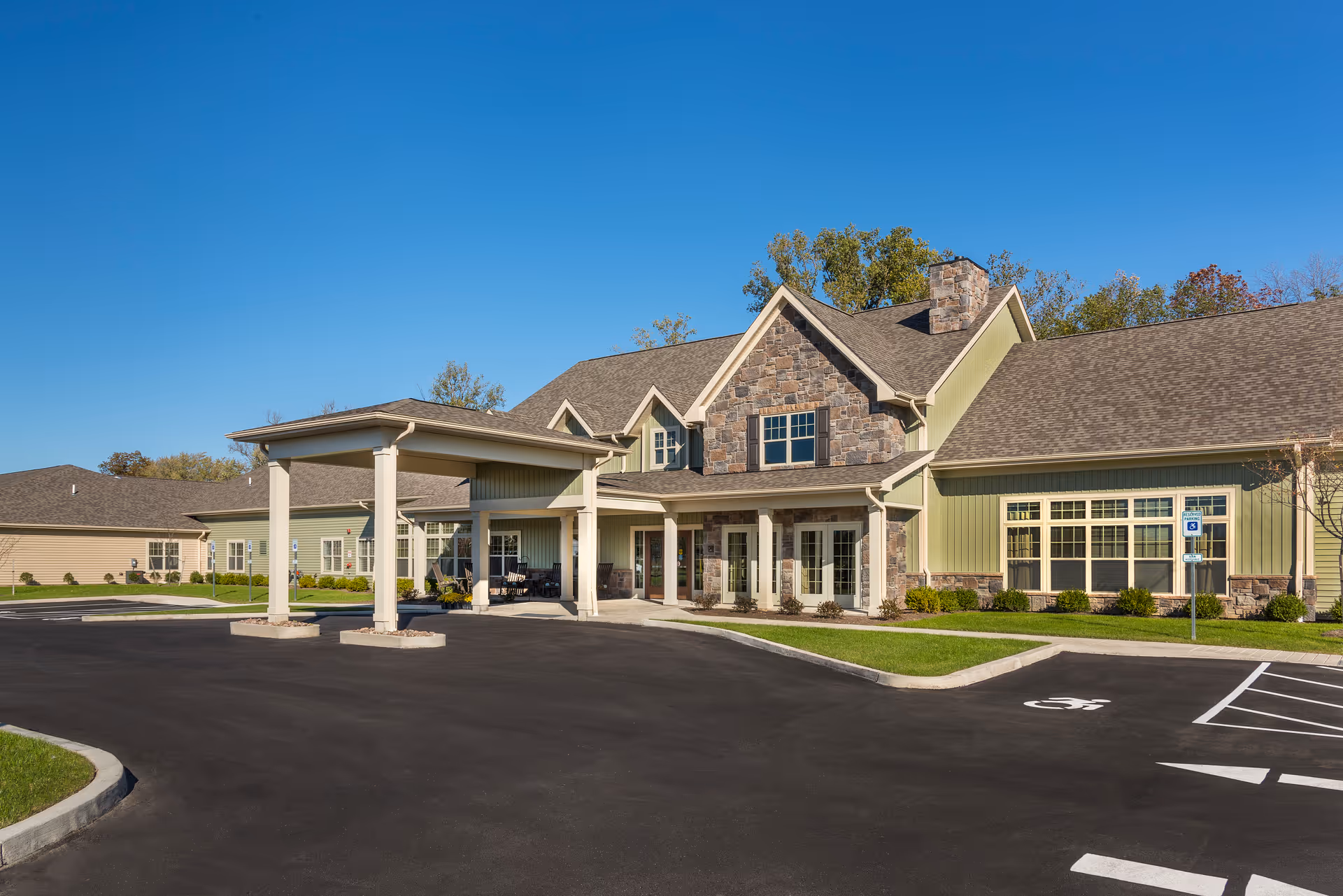 Front exterior of a senior living community building with stone accents, a covered entrance/portico and adjacent parking lot under a clear blue sky.