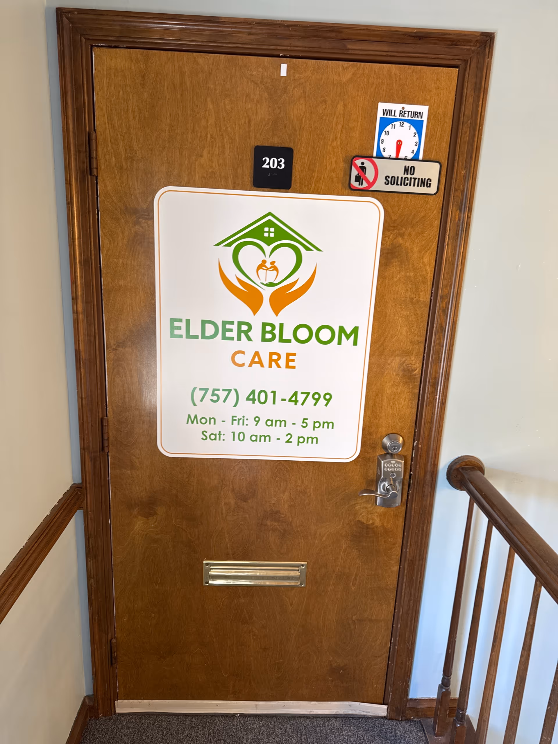 Wooden interior door with Elder Bloom Care sign, room number 203, no soliciting, staircase railing nearby.