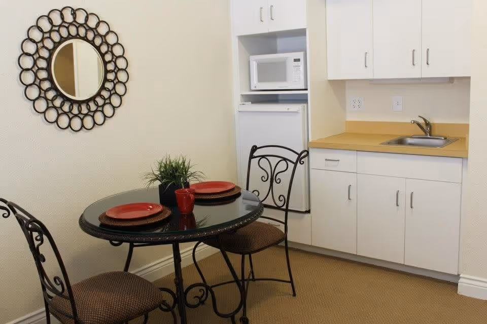 A small dining area with a round glass-top table set with two red plates and a red mug, accompanied by two wrought iron chairs with cushioned seats. The area is adjacent to a kitchenette featuring white cabinets, a small sink, a microwave, and a mini refrigerator. A decorative circular mirror with a black frame hangs on the wall above the table.