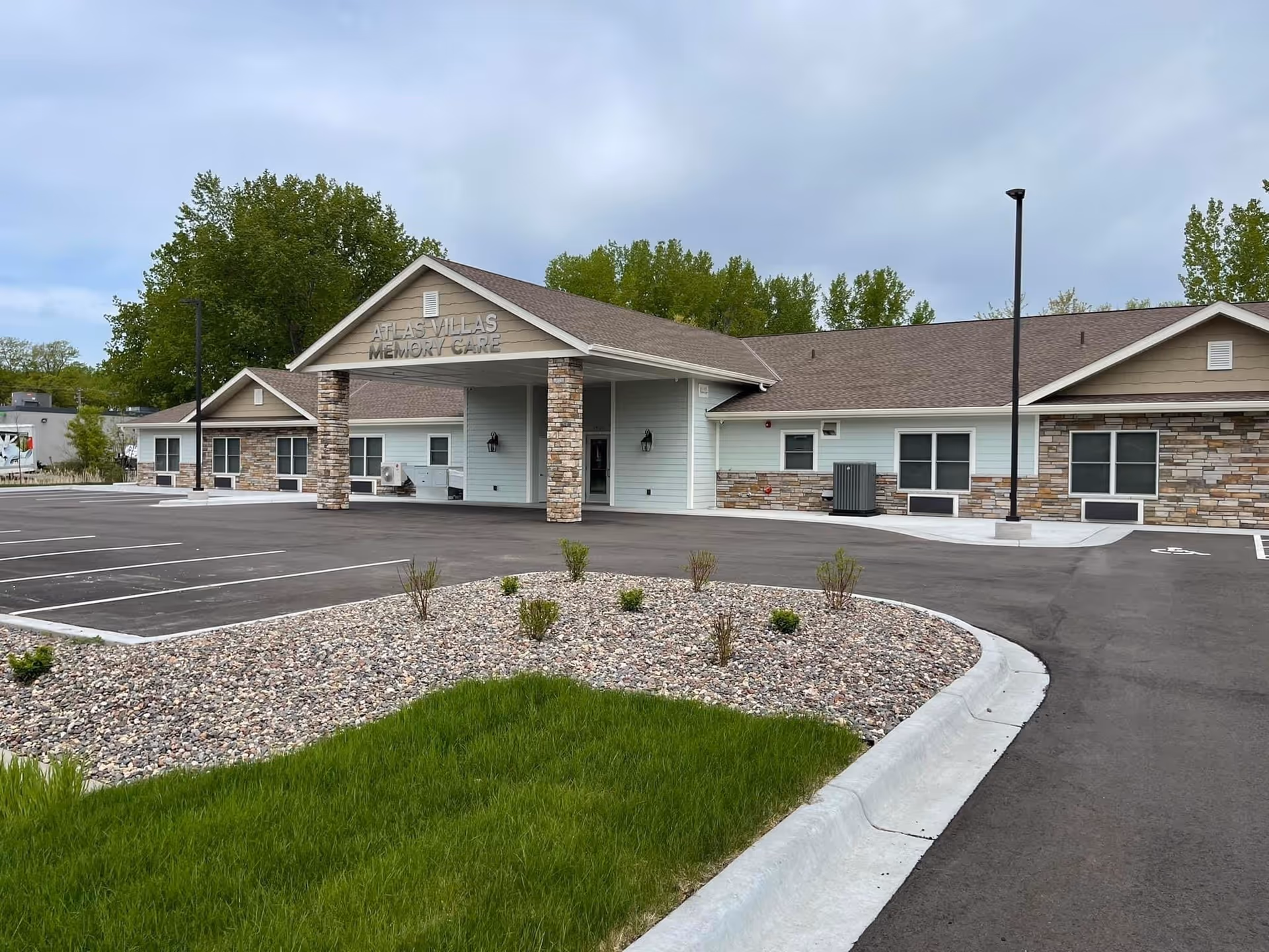 Exterior view of Atlas Villas Memory Care building with a covered entrance supported by stone pillars, surrounded by a parking lot and landscaped areas with grass and small plants under a cloudy sky.