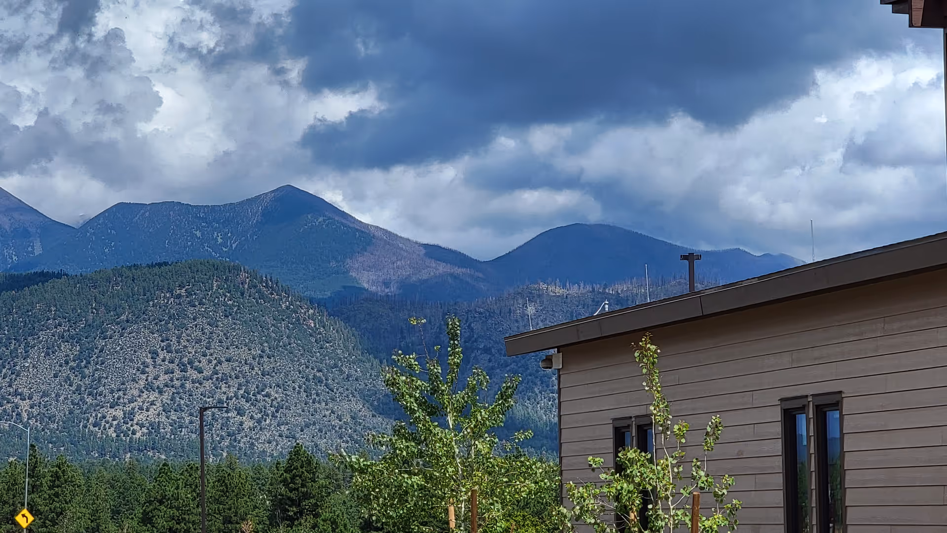 Exterior view of a facility with a brown wall, trees, and distant mountains beneath cloudy skies.
