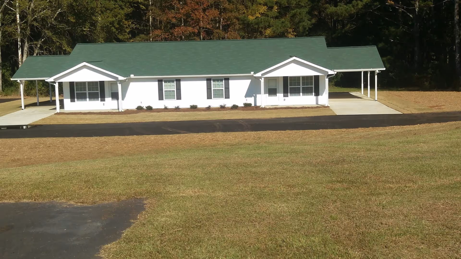 Single-story white building with a green roof, multiple windows, and two covered carports on either side, surrounded by grass and trees in the background.
