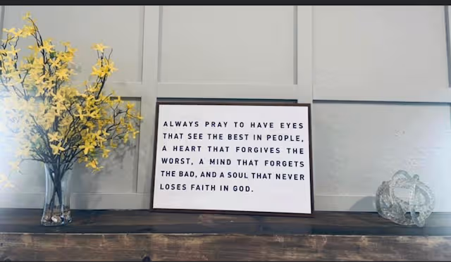 A framed sign with an inspirational quote sits on a wooden shelf beside yellow flowers.