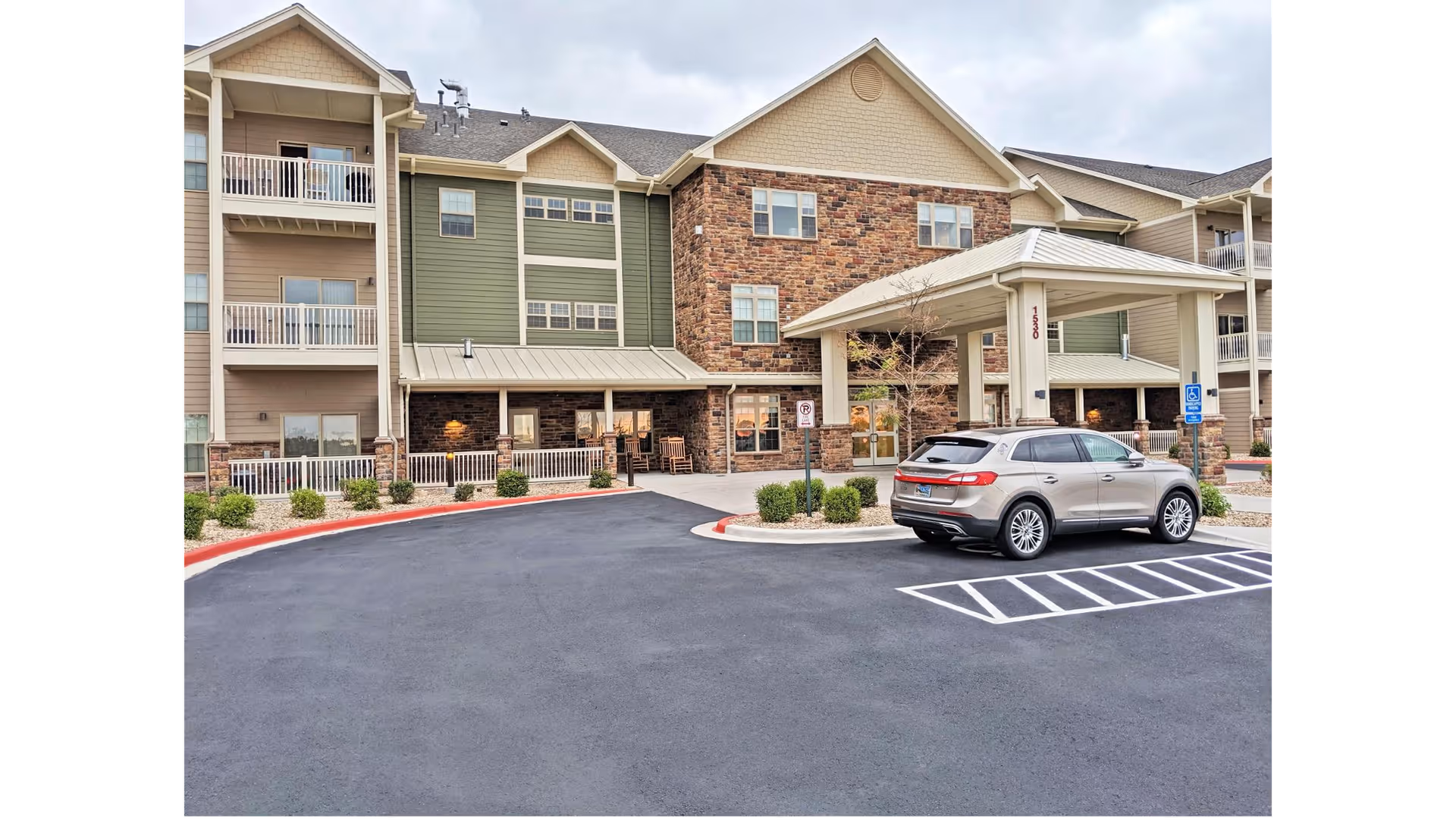 Exterior view of a multi-story retirement community building with a covered entrance and a parked silver SUV in front. The building features a combination of stone and siding with balconies and windows. The area is paved with parking spaces and some landscaping with bushes.