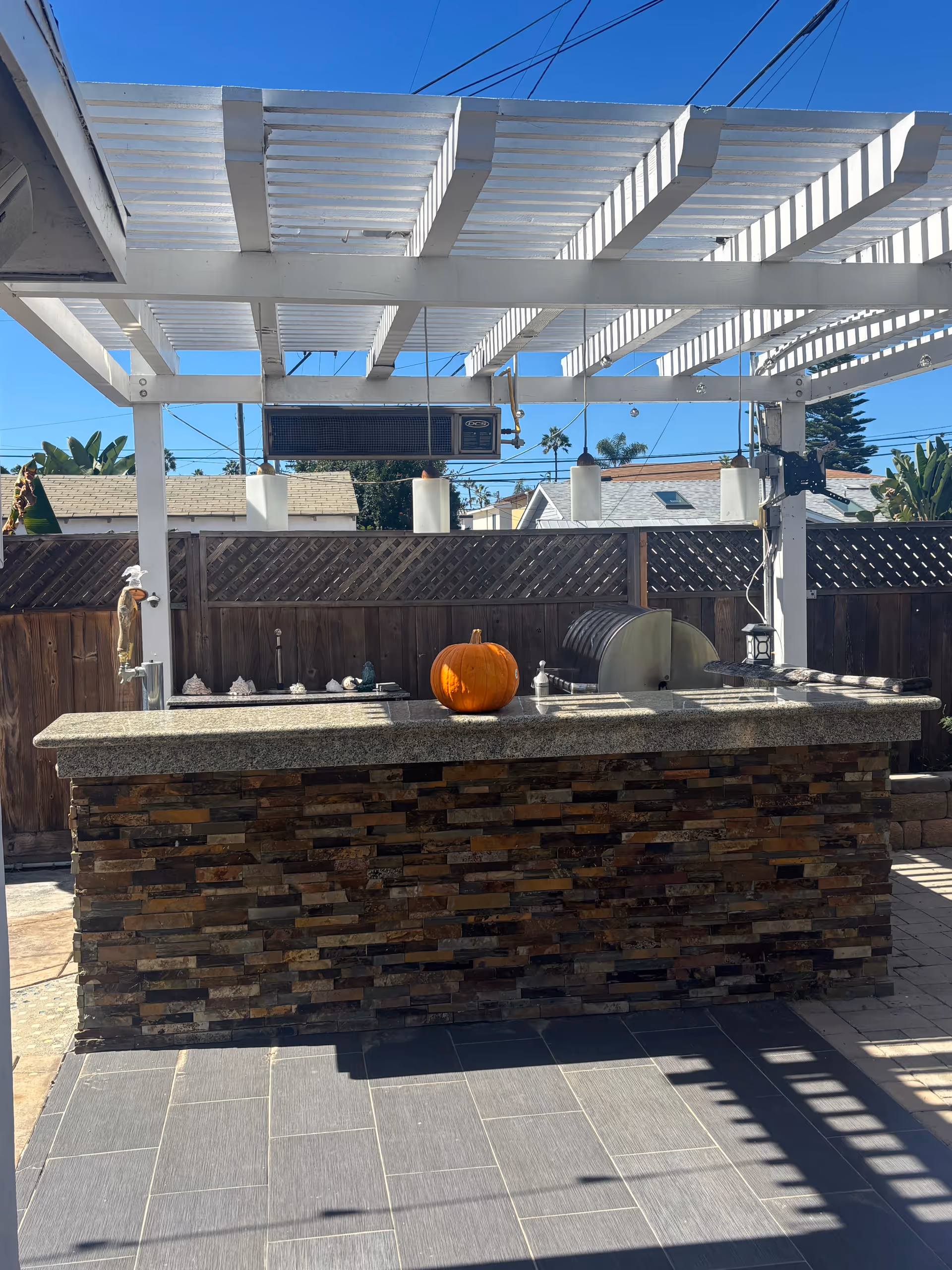 Outdoor kitchen under a pergola with a stone island, grill, and pumpkin