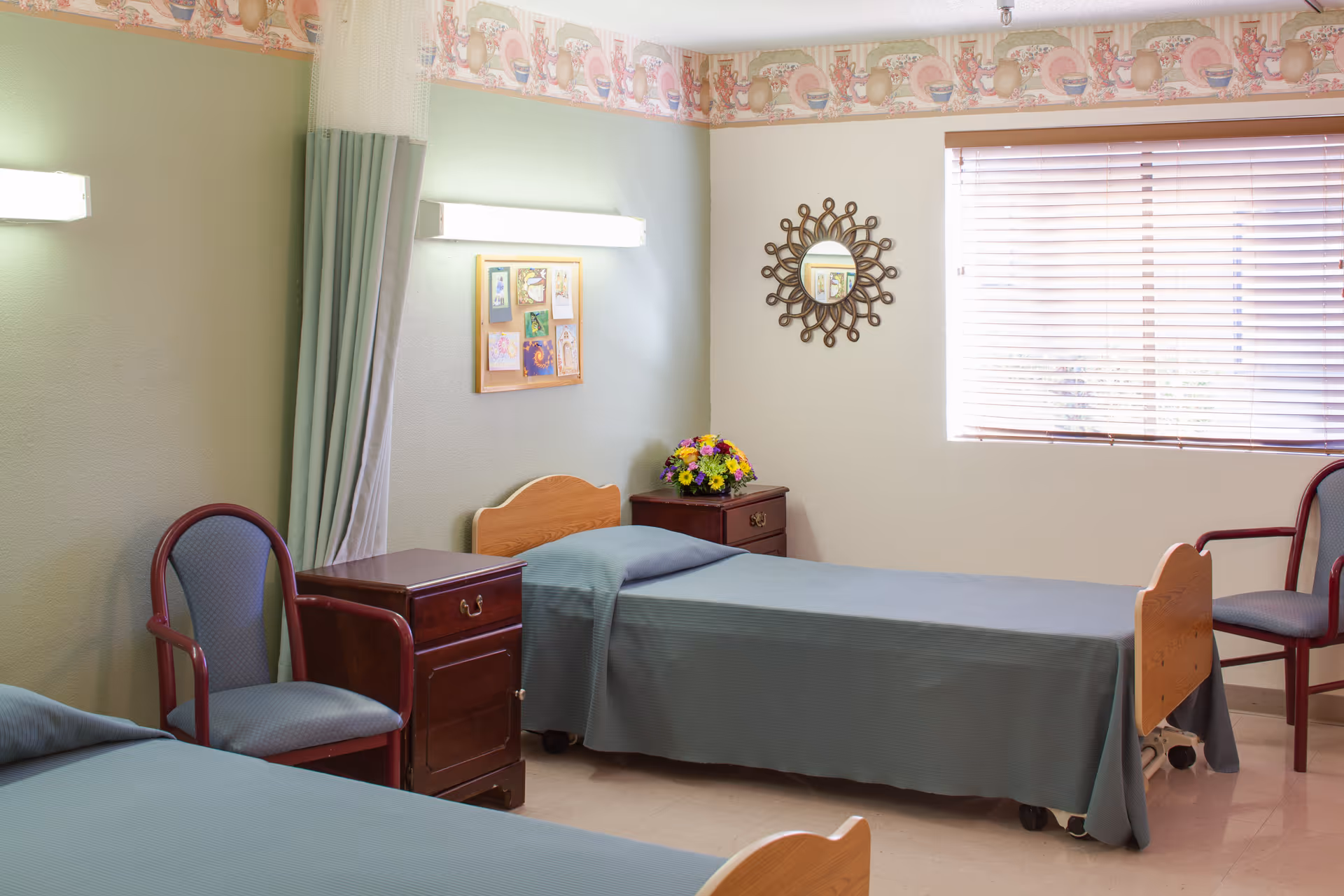 A simple shared bedroom with two single beds, wooden nightstands, chairs, and a window with blinds.