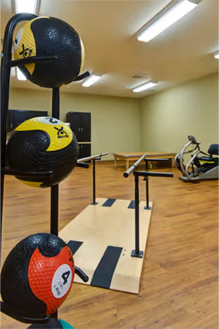 Indoor fitness room with wooden flooring featuring a rack of three medicine balls in the foreground, parallel bars for walking exercises in the center, and an exercise bike in the background under fluorescent ceiling lights.