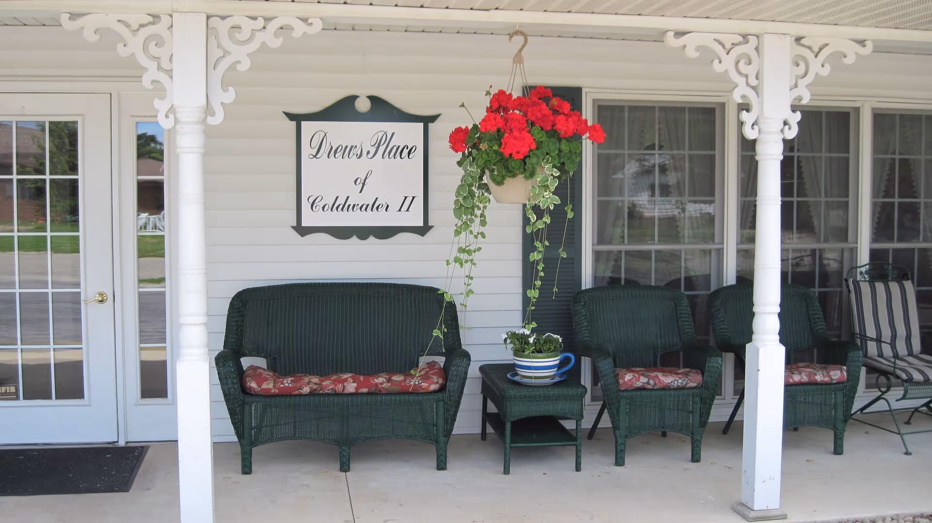 Front porch area of Drews Place of Coldwater II featuring white siding, decorative white columns, a hanging pot of red flowers, a green wicker loveseat and chairs with floral cushions, a small green wicker table with a striped flower pot, and a glass door with white framing.