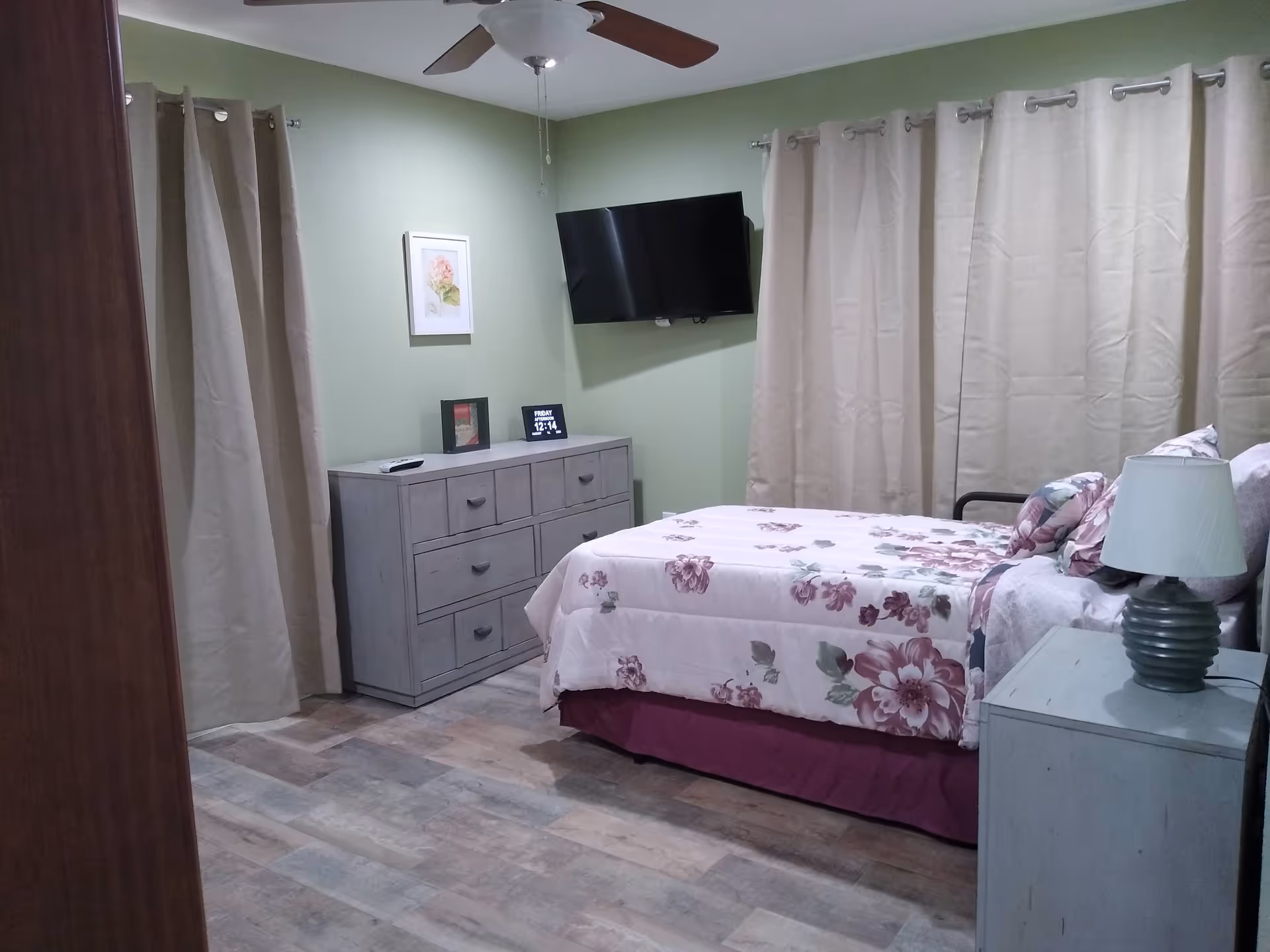 A bedroom with floral bedding, dresser, wall-mounted TV, beige curtains, and a ceiling fan.