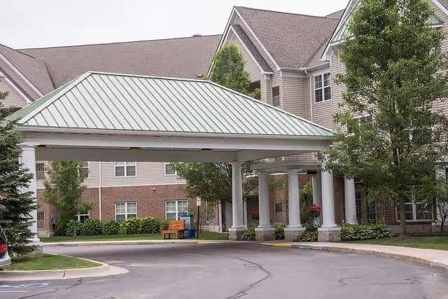 Exterior view of a senior living facility with a covered entrance supported by white columns. The building has beige siding with brick accents and multiple windows. There are trees and shrubs around the entrance, and a bench is visible near the building.
