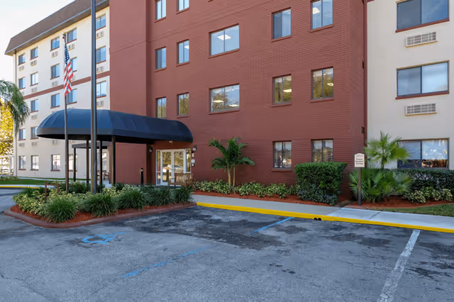 Exterior view of a multi-story senior living facility building with a red brick section and beige sections. There is a black awning over the entrance door, an American flag on a flagpole, and landscaped greenery around the entrance. The parking area in front includes a handicapped parking space.