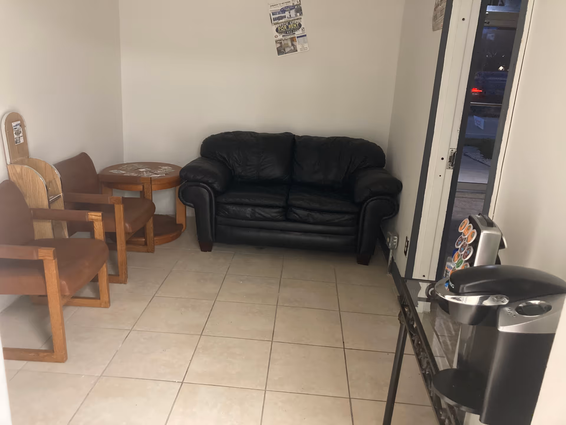 Small waiting area with a black leather loveseat, wooden chairs, round table, and a vending machine.