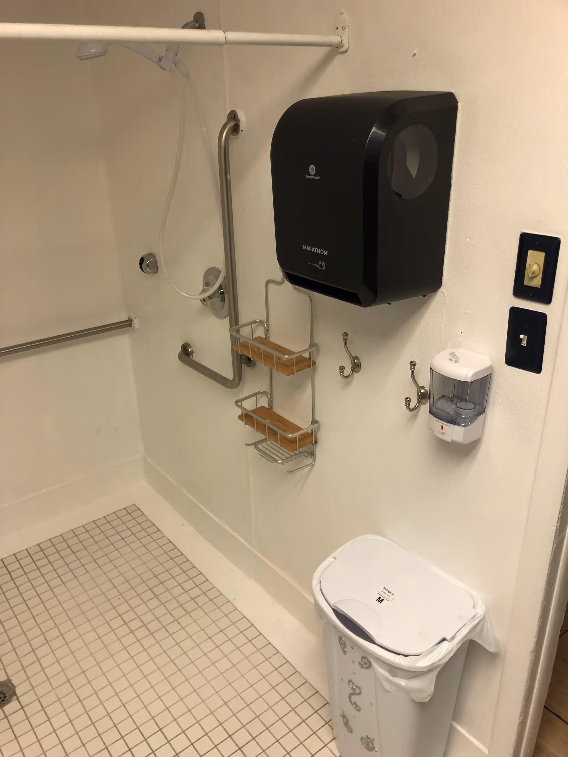 Accessible bathroom with a shower, grab bars, wooden shelves, and wall-mounted soap dispenser.