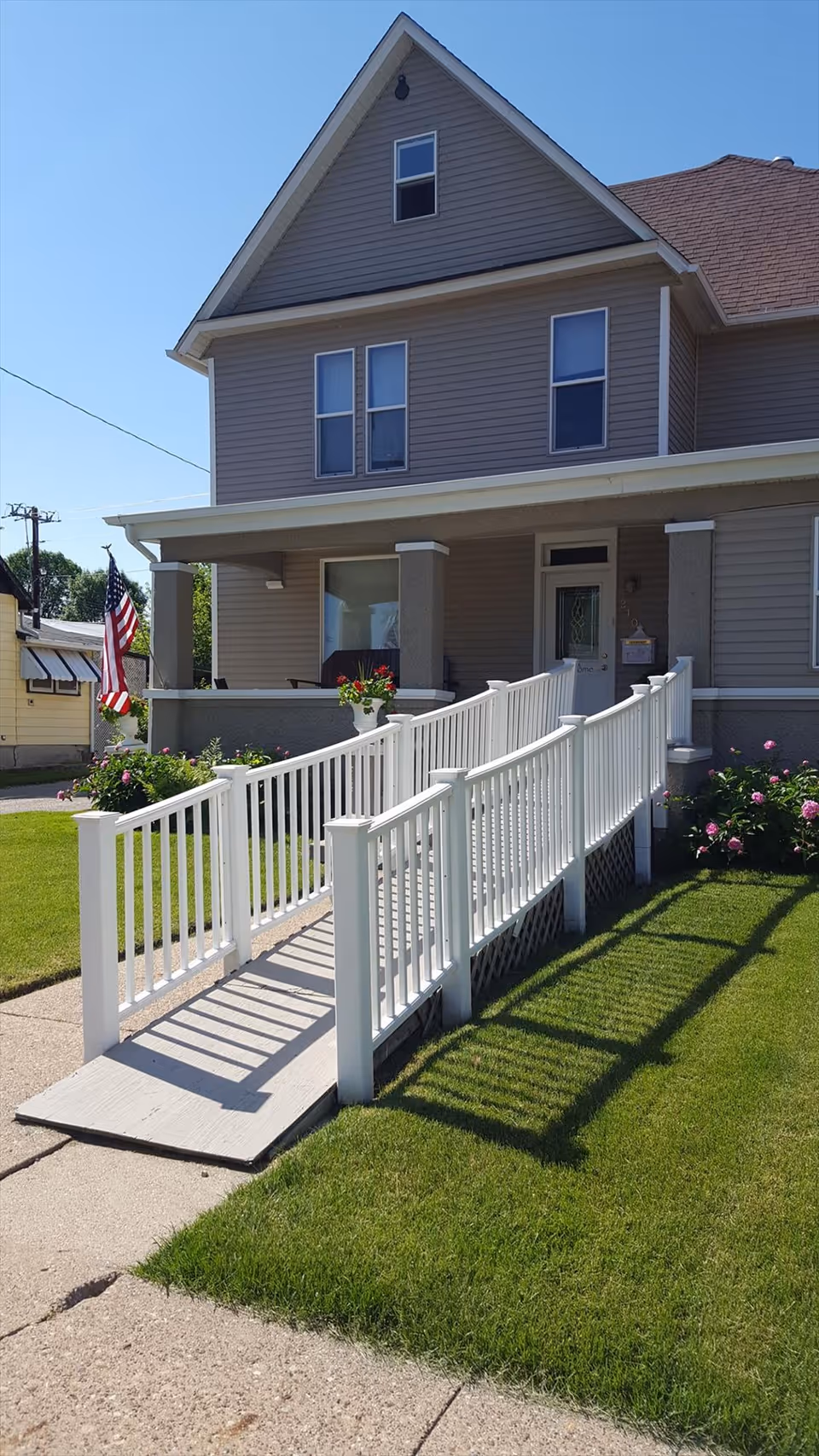 Exterior view of a two-story house with beige siding and a brown roof. The house has a covered front porch with a white wheelchair ramp leading up to the entrance. There are green lawns and flower bushes on either side of the ramp, and an American flag is displayed near the porch.