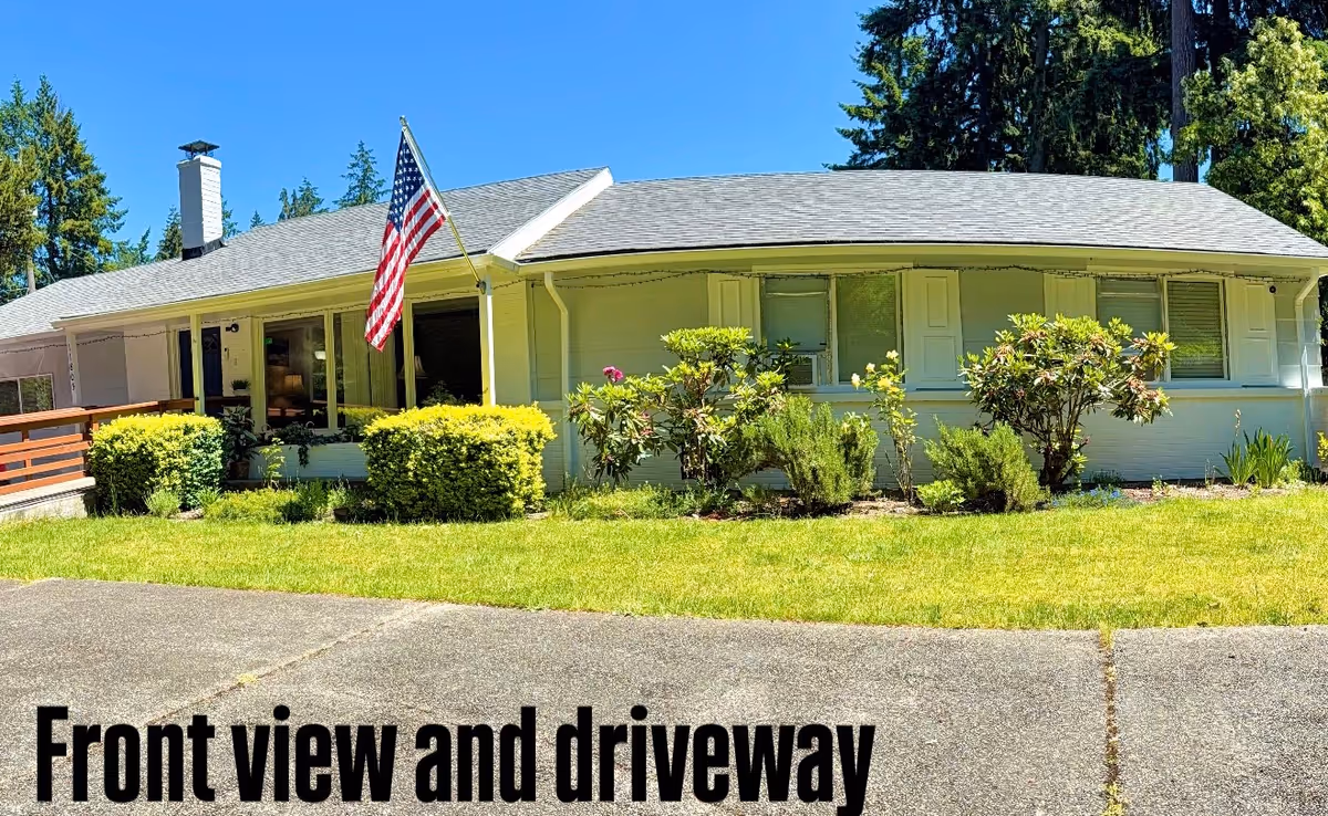 A single-story light-blue house with an American flag, shrubs, and a paved driveway.