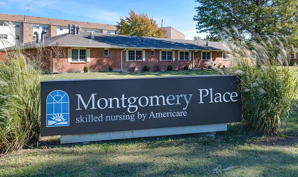 Outdoor view of a single-story brick building with multiple windows, behind a large sign that reads 'Montgomery Place skilled nursing by Americare'. The sign is black with white text and a blue logo on the left side. There is grass and tall ornamental grass plants around the sign, and trees in the background under a clear blue sky.