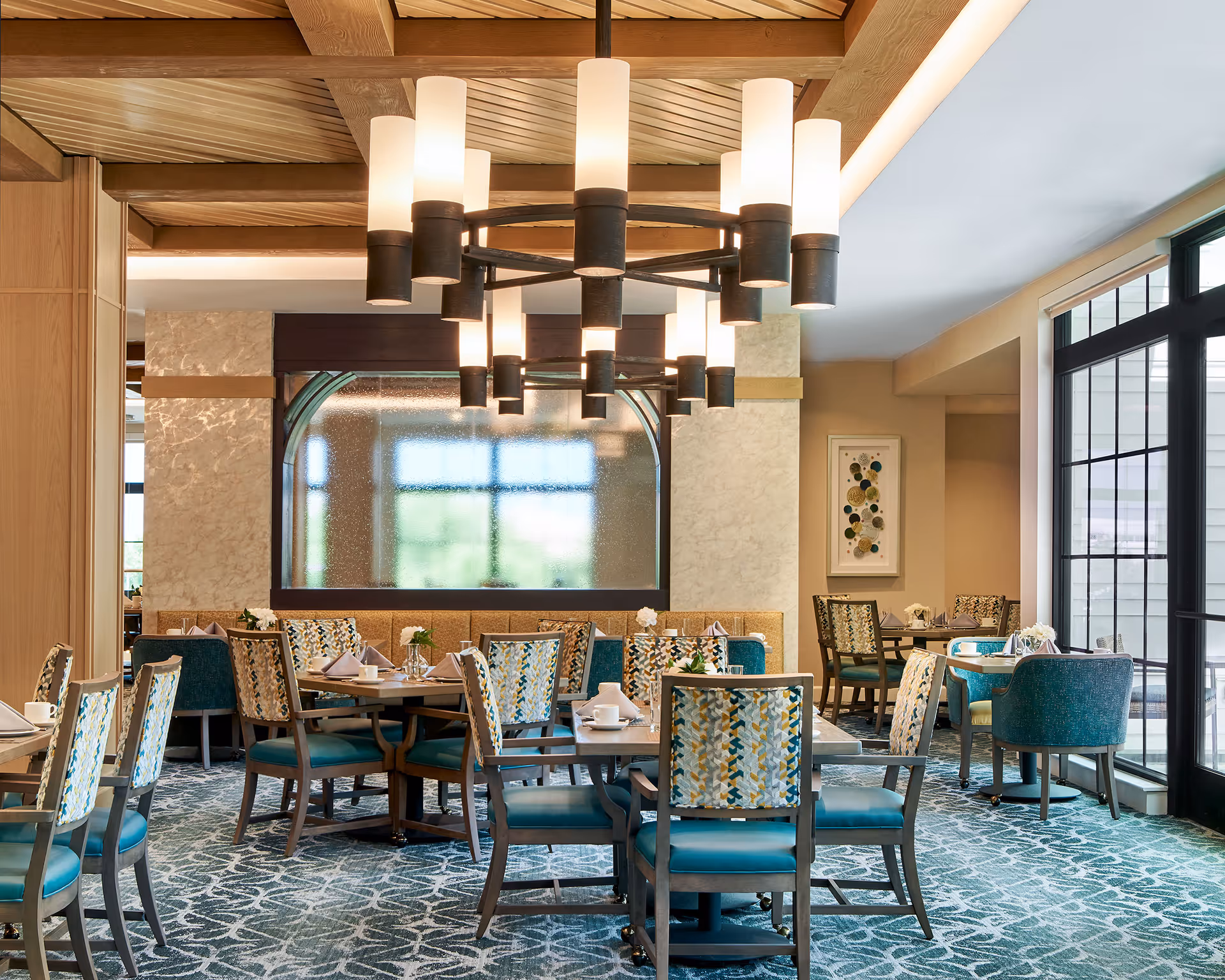 A bright and elegant dining room with multiple tables and chairs arranged neatly. The chairs have patterned upholstery and teal cushions. A large modern chandelier with cylindrical lights hangs from a wooden ceiling. There is a large window on the right side allowing natural light to fill the room, and a decorative wall piece is visible in the background.