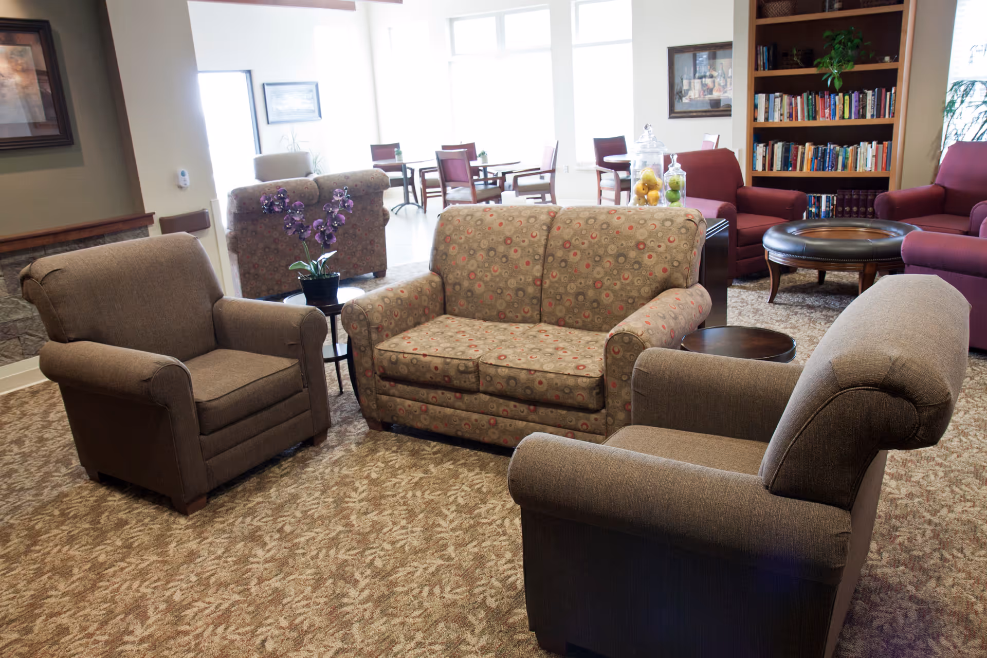 A cozy living room area with a patterned loveseat and two brown armchairs arranged around a small round table. In the background, there are more chairs, tables, a bookshelf filled with books, and large windows letting in natural light.