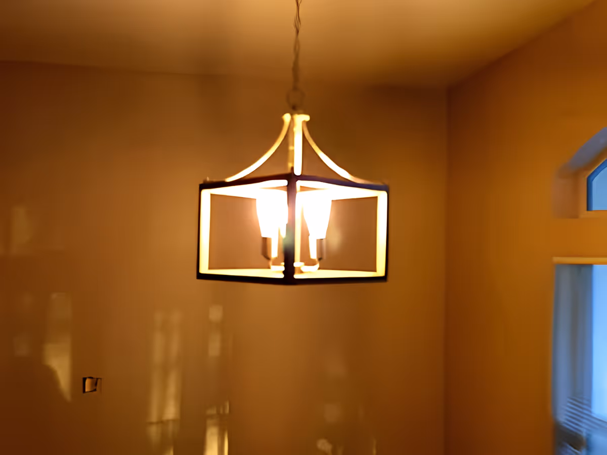 A warm-lit modern square chandelier hangs in a beige interior room with a small arched window visible.