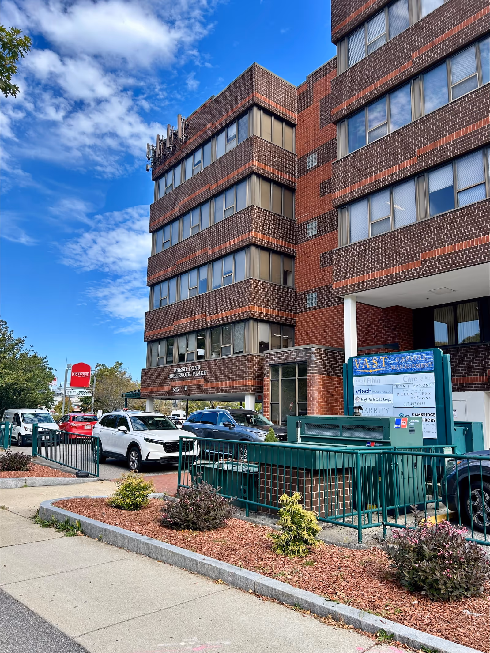 Brick, multi-story senior living building exterior with parked cars, landscaping, and blue sky.