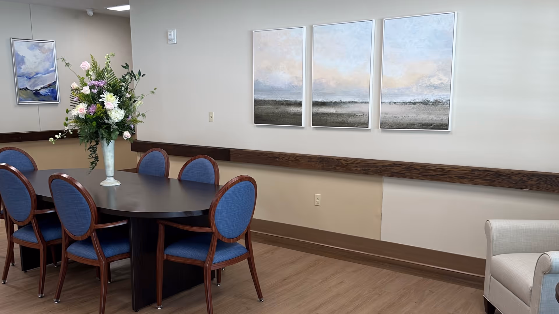 Dining area with a dark round table, blue chairs, floral centerpiece, and wall art.