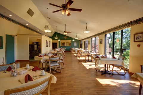 A bright dining room with multiple tables set with white tablecloths, red napkins, and cups. The room has large windows on one side letting in natural light and offering a view of greenery outside. Ceiling fans and hanging lights are visible, and the floor is wooden.