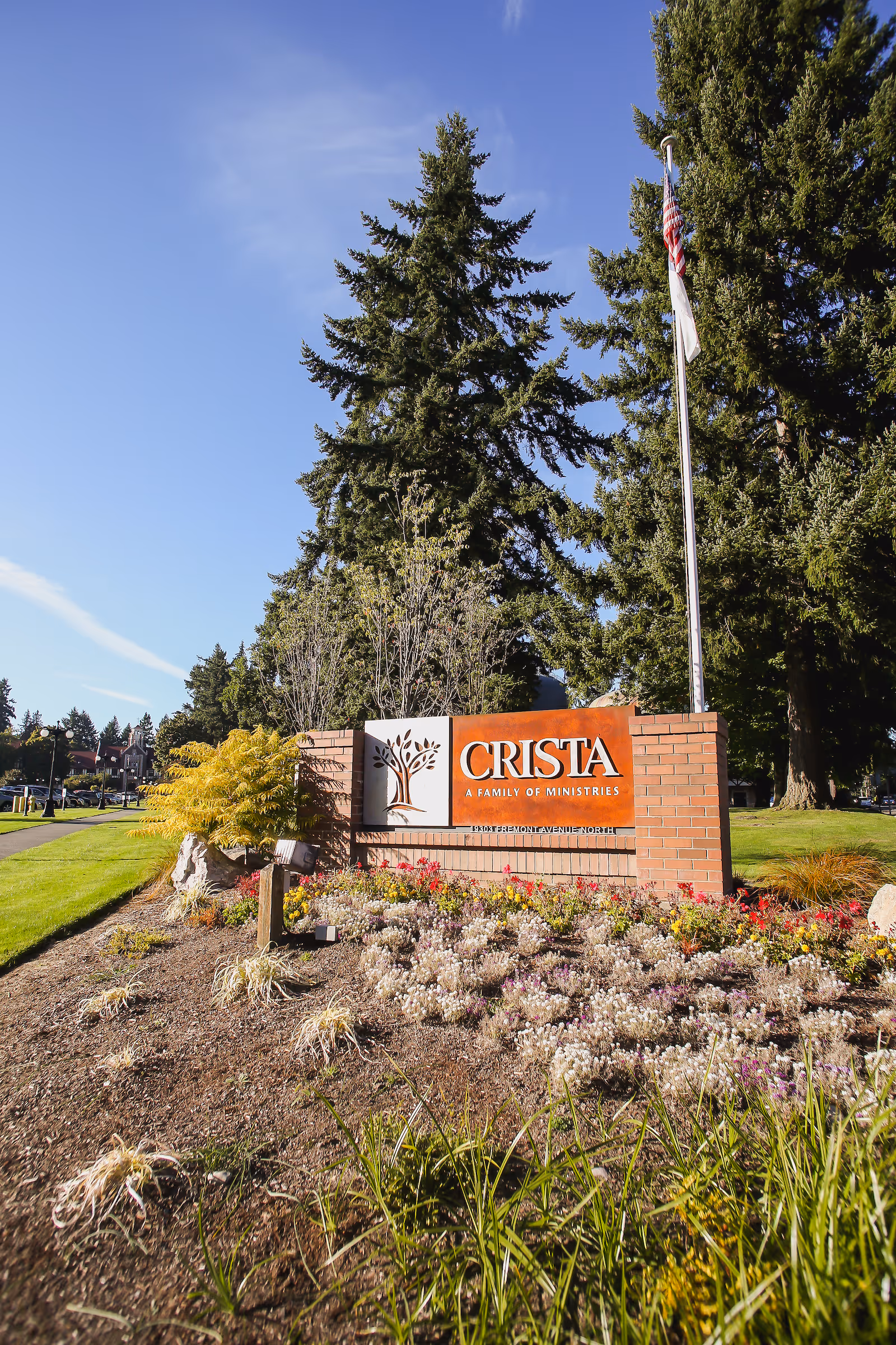 Brick entrance sign reading 'CRISTA' set in landscaped grounds with flowers, tall trees, and a flagpole.