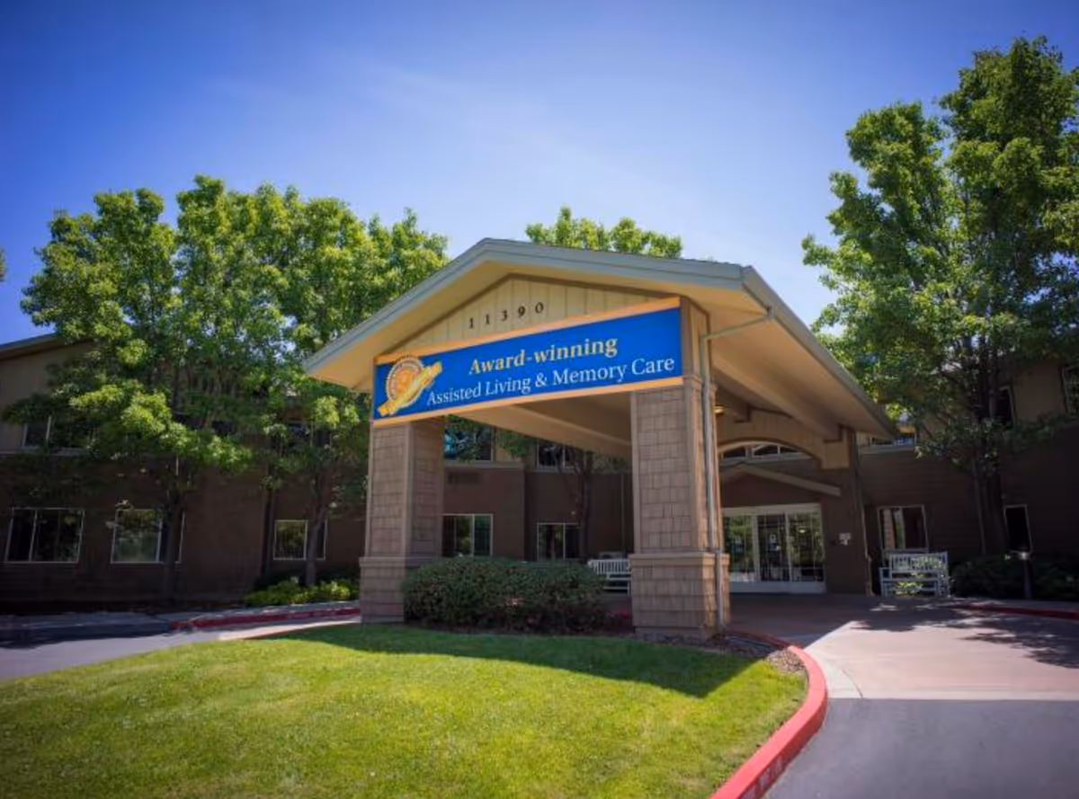 Entrance canopy of a senior living facility with a blue sign reading "Award-winning Assisted Living & Memory Care" in front of a brick building and trees.