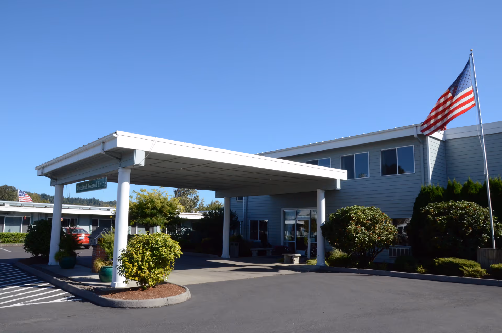 Exterior view of Woodland Care Center & Assisted Living building with a covered entrance, landscaped bushes, an American flag on a flagpole, and a clear blue sky.