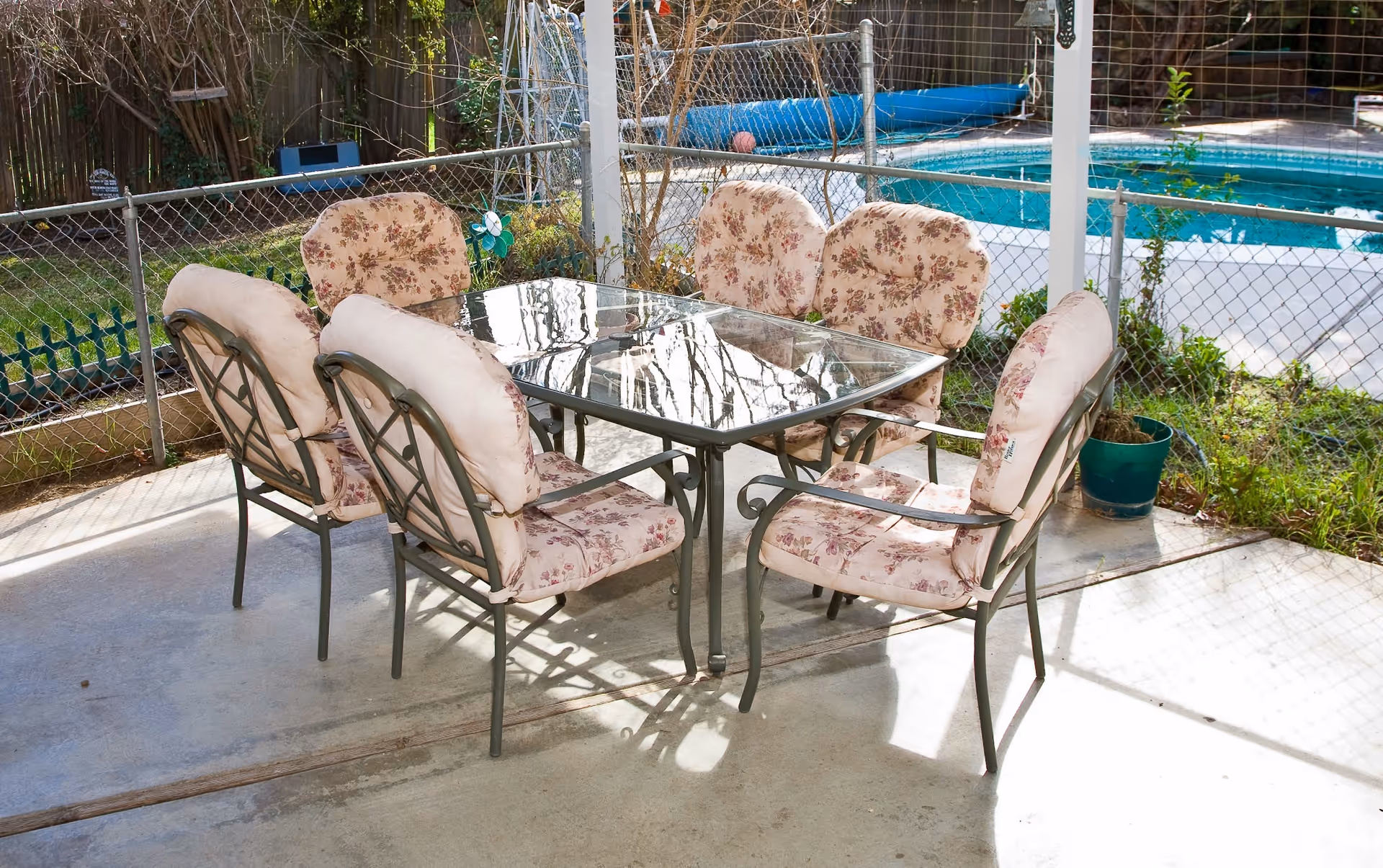 Outdoor patio area with a glass-top table surrounded by six cushioned chairs with floral patterns. The patio is enclosed by a chain-link fence, and a swimming pool is visible in the background. There is a potted plant on the patio floor near the fence.