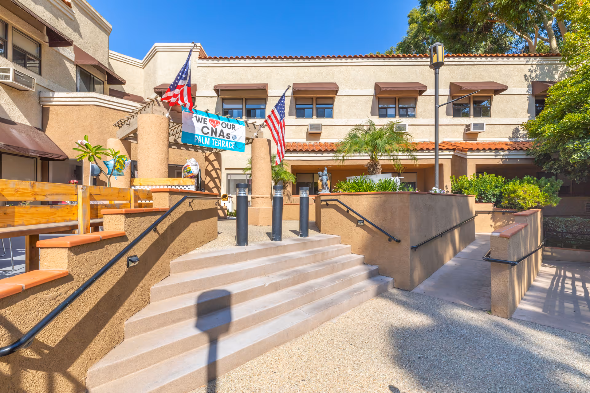 Entrance to a senior living facility with stairs and a wheelchair ramp leading to a courtyard decorated with American flags and a banner that reads 'We ♥ Our CNAs Palm Terrace'.