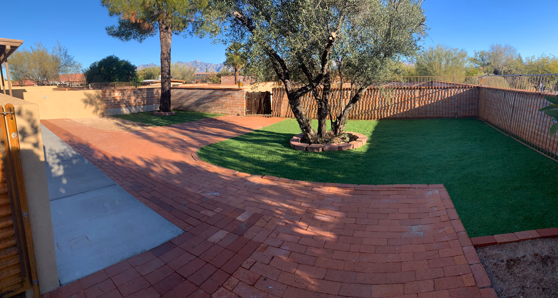 Outdoor courtyard area with red brick paving and green artificial grass patches. A tree is planted in the center of one grassy area, surrounded by a circular brick border. The space is enclosed by brick and metal fencing, with desert vegetation and mountains visible in the background under a clear blue sky.