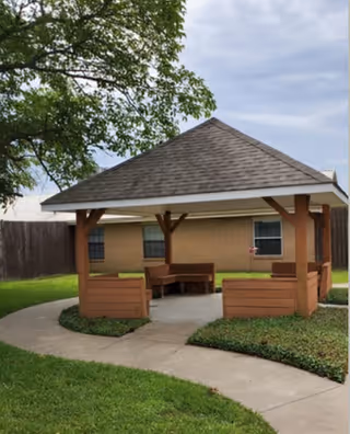A wooden gazebo with a shingled roof situated on a concrete pathway surrounded by green grass and landscaping. The gazebo has built-in wooden benches and is located near a beige building with windows and a wooden fence in the background under a cloudy sky.