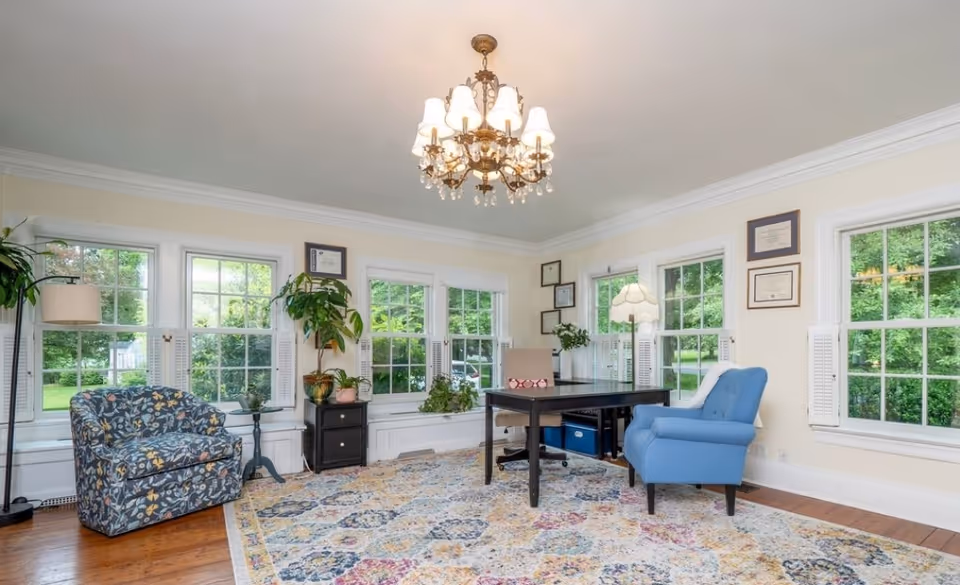 Bright living room with chandelier, large windows, plants, desk, and seating.